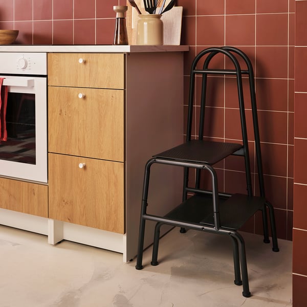 Kitchen scene: modern brown-tiled wall, light wooden cabinet with oven, grey metal 2-step stool with black steps and handrail.