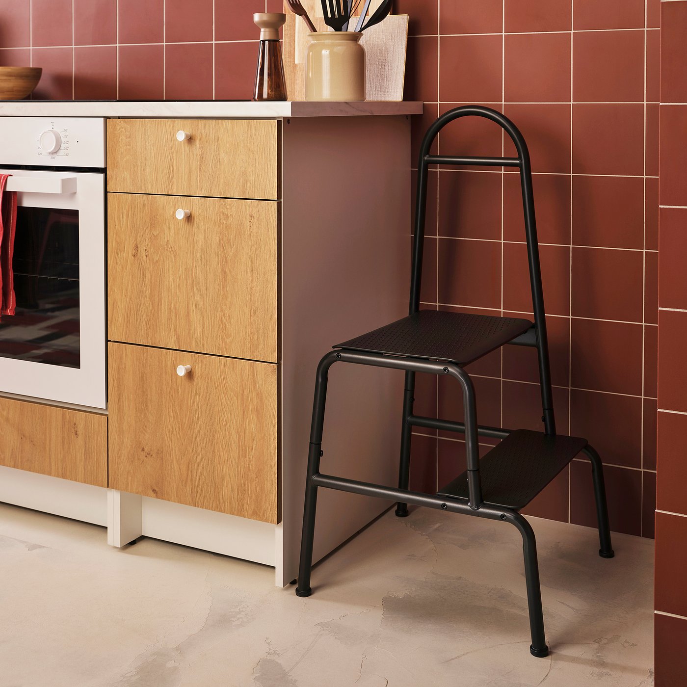 Kitchen with black BÄSINGEN 2-step stool, dark grey metal frame, wooden cabinets, and red tiles.