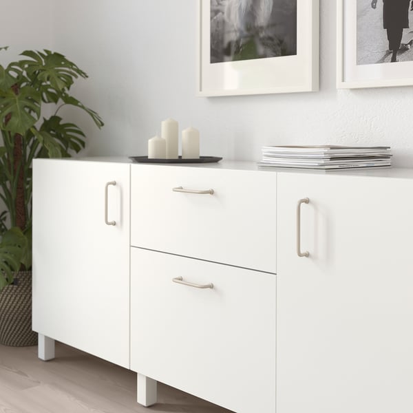 Modern white BAGGANÄS cabinet with sleek handles, medium-sized, holding candles and magazines beneath framed photos and a tall plant.