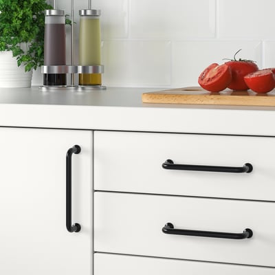 Modern kitchen drawer handles in black, BAGGANÄS style, with visible screws, set against a countertop displaying tomatoes and bottles.