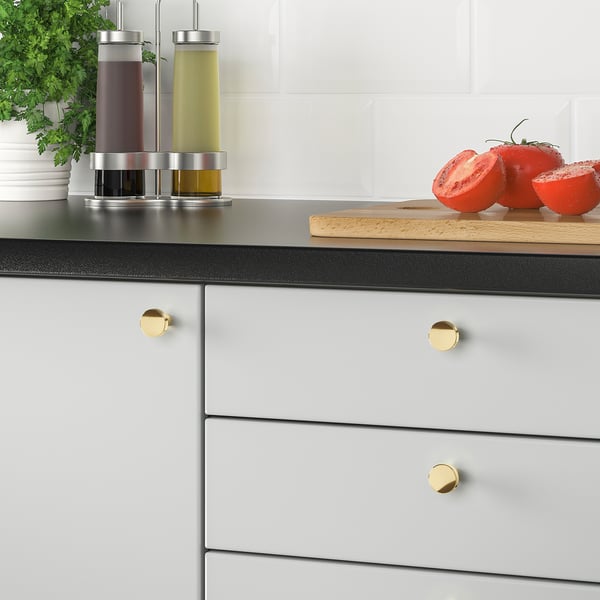 Modern kitchen with BAGGANÄS brass cabinet knobs. Black countertop holds tomatoes and herbs.