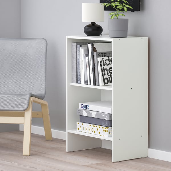 White wall-mounted bookcase with open shelves holds books and decorative items, including a lamp and a plant.