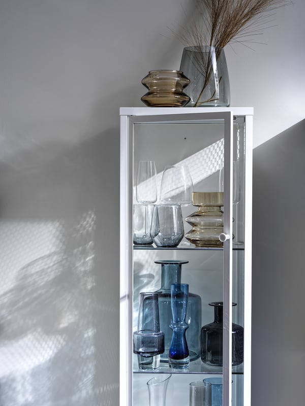 White BAGGEBO display cabinet with blue and clear glass vases and wine glasses.