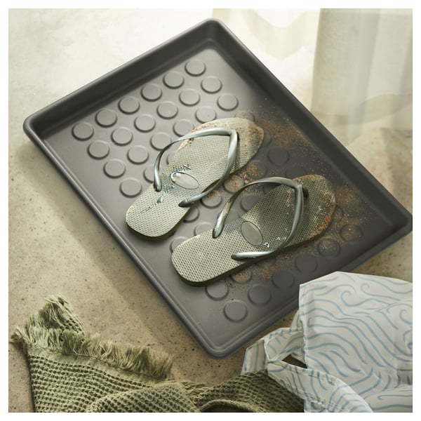 Black BAGGMUCK mat with flip flops, textured circles, on beige floor.