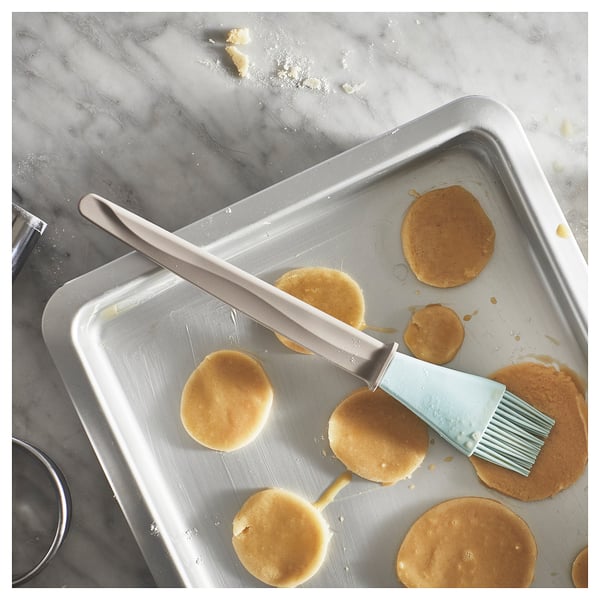Silver baking brush on tray with baked goods. Brush has blue handle and silicone bristles. Tray is metallic with round baked items.