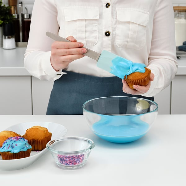 Person icing a muffin with a blue spatula, using a clear bowl for icing.