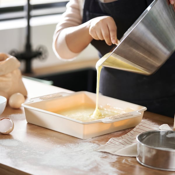 Person pouring liquid batter into cake pan on wooden table.