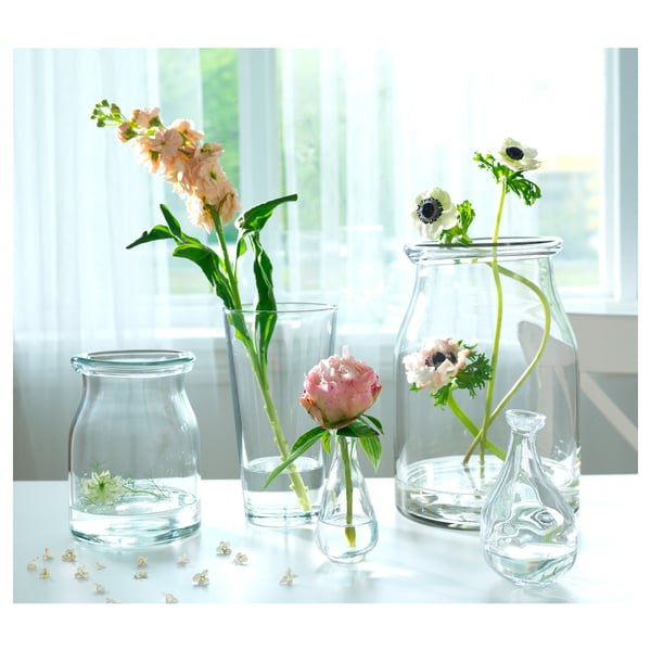 Five clear glass vases of varying shapes hold fresh flowers, placed by a window.