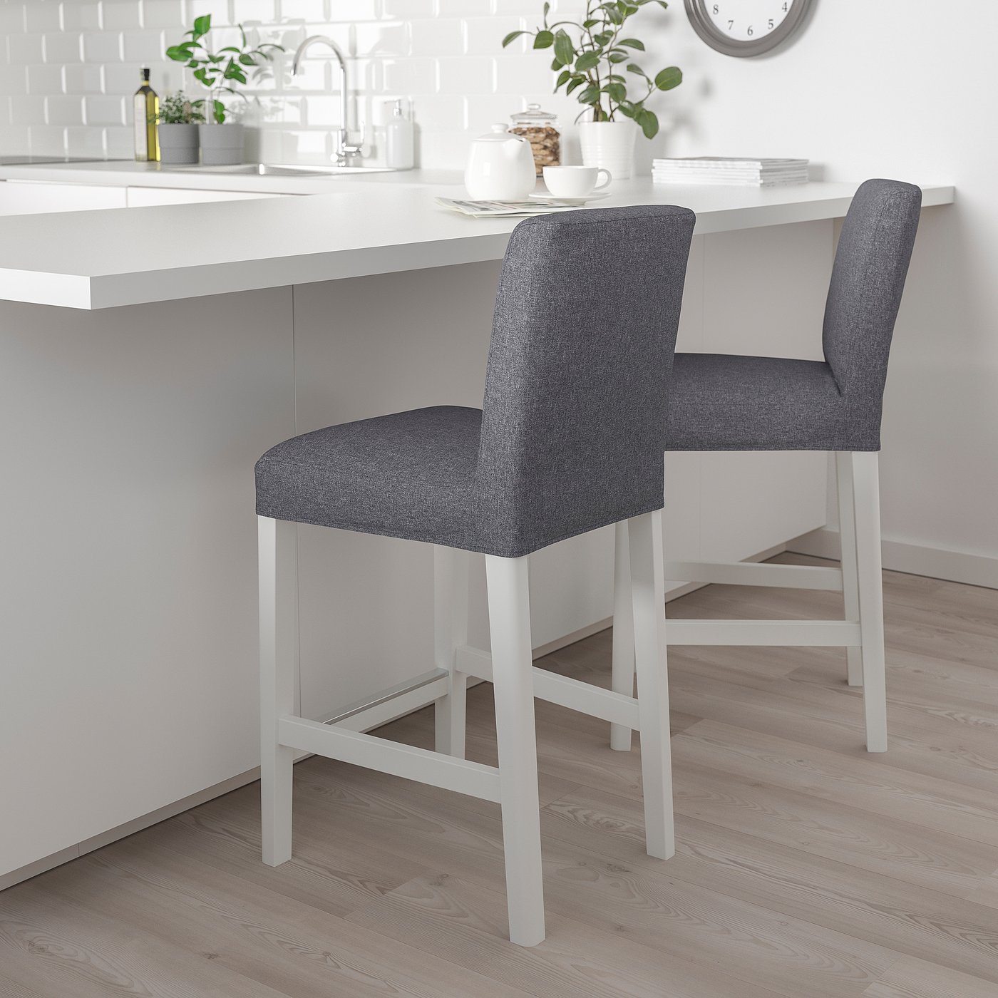 Two grey upholstered bar stools with white legs at a white kitchen island. The stools are taller than the island, with cushioned seats and backrests.