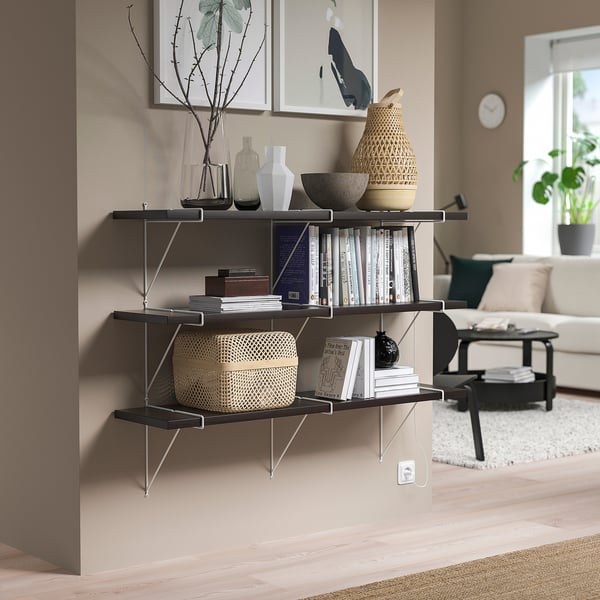 Black BERGSHULT wall shelf, holding various items, stands tall, filling a corner beside a beige wall, with a modern, minimalist design.
