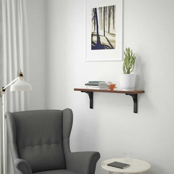 Minimalist living space with walnut shelf, books, cactus, chair, table, and forest picture. Lit by natural light and wall lamp.