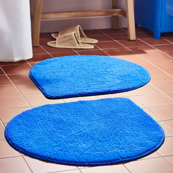 Two bright blue BERGVATTNET bath mats, oval-shaped, soft, on brown tiles, larger than a pair of slippers nearby.