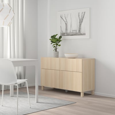 Minimalist room with BESTÅ oak cabinet against white wall, featuring doors, plant, bowl, and framed trees picture near desk and chair.