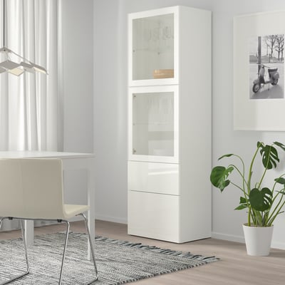 White BESTÅ cabinet stands tall, housing glasses behind clear doors, near a desk and plant.