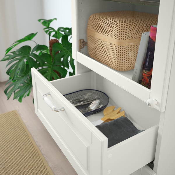 White BESTÅ unit with open drawer showing cutlery tray and folded napkins, atop shelf with basket and books.