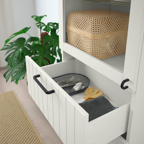 White BESTÅ cabinet with open drawer showing utensils and a wicker basket on upper shelf. Green plant nearby.