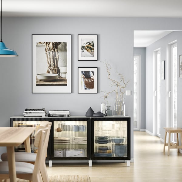 Modern dining area with light wood table and chairs, black and clear sideboard, grey wall, three framed photos, blue light fixture.