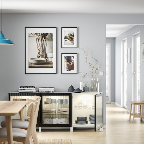 Modern living room with light grey walls, wooden floor, and dining area. Black and white furniture, framed art, vases, and books.