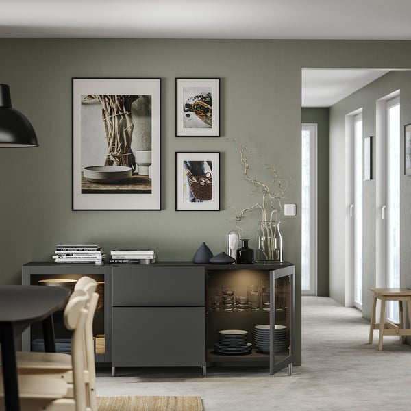 Modern dining room with grey BESTÅ sideboard, wooden chairs, and black pendant light. Green walls, framed art, and glassware displayed.