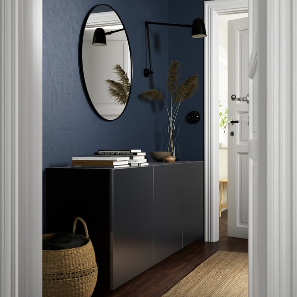 Modern entryway with BESTÅ grey cabinet, round mirror, dark walls. Basket and books on cabinet, door slightly open.