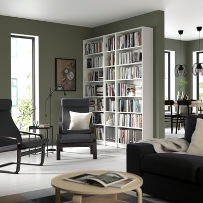Modular white bookshelf filled with books, between green walls, with black furniture, creating a cosy reading nook.