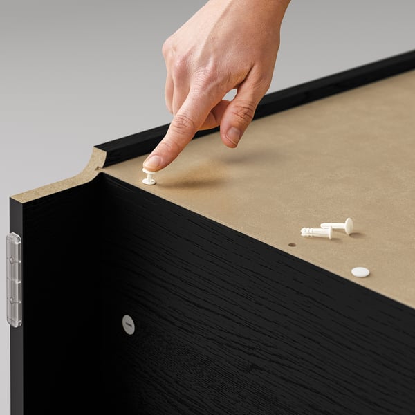A hand placing a white peg into a black BILLY bookcases corkboard top, fixing it with two other pegs nearby.