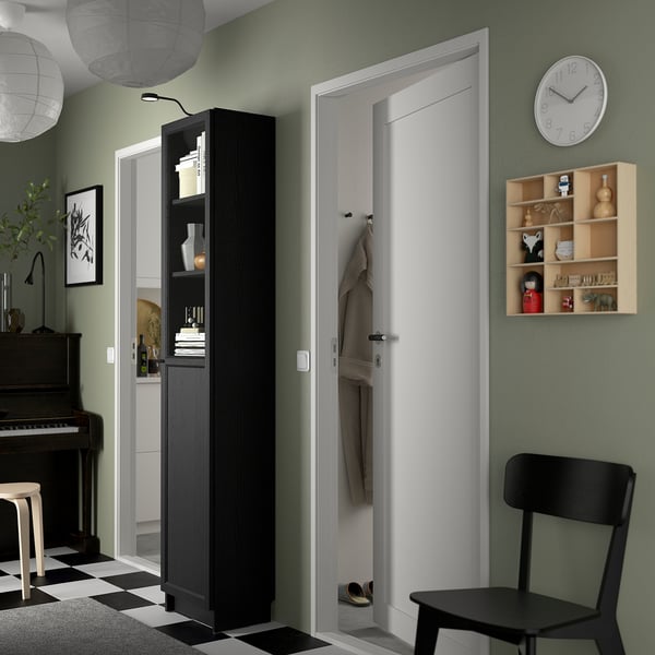 BILLY bookcase in black beside door, filled with items, panel door, chair, wooden shelf on green wall.