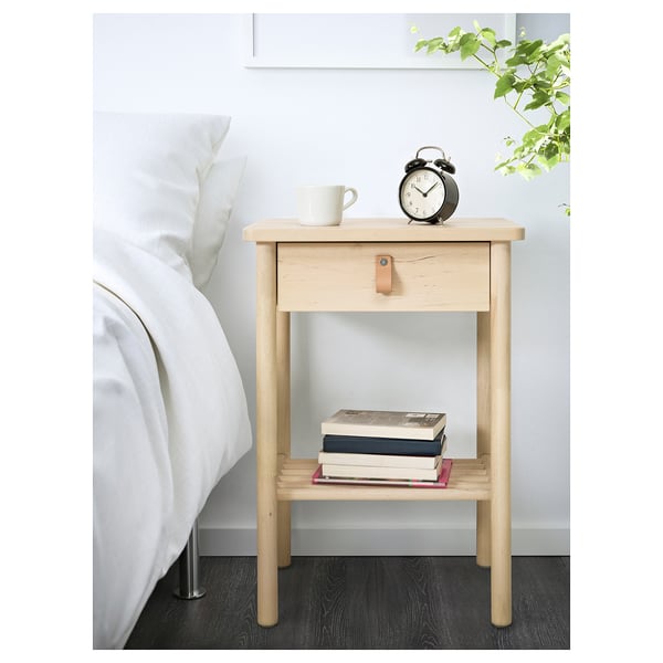 Wooden bedside table with drawer and shelf, holding books, a mug, and an alarm clock next to a white bed.