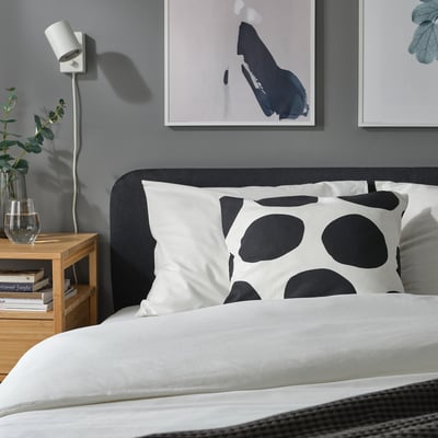Bedroom with white pillow with black dots, grey headboard, minimalist decor, abstract art, and bedside table with vase and books.