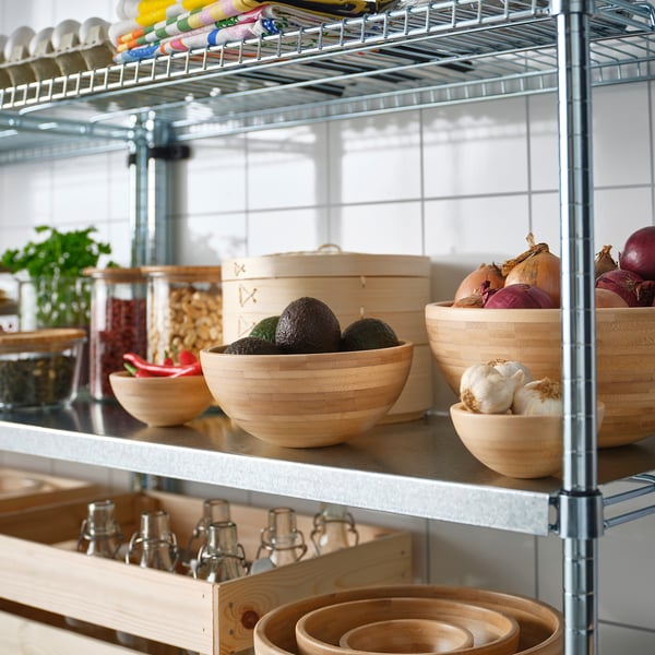 A kitchen shelf with wooden bowls, one containing avocados, another with onions and garlic. Above, jars and spice bottles are neatly organised.
