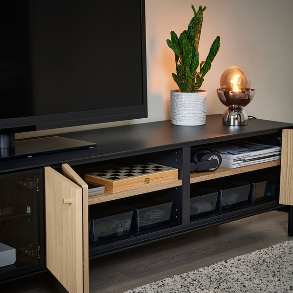 Modern tv stand with glass doors, open compartment showing chess set, plant, and lamp. Black and wood combo, stylish and organised.
