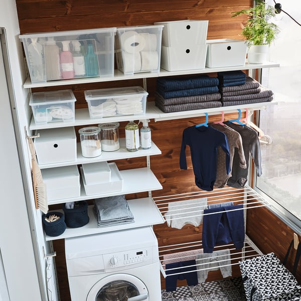 Organised laundry room with white BOAXEL shelves storing clothes, detergents, and towels, plus a drying rack hanging garments by the window.