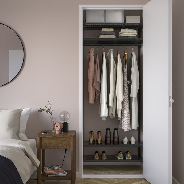 Closet with neutral clothes on black BOAXEL shelves. Bedside table with clock, lamp, and plant. Dark shelves contrast pink wall.