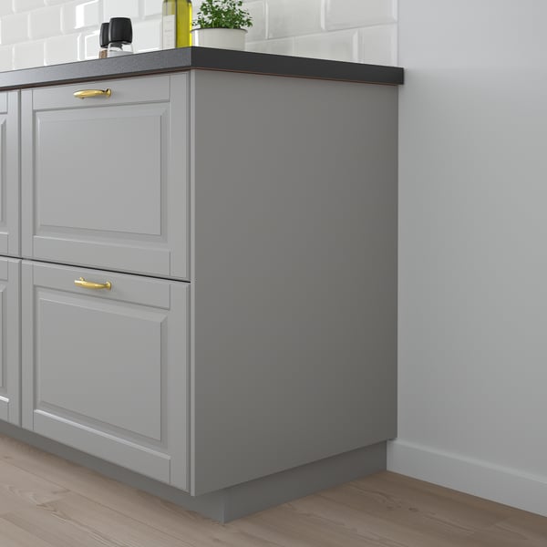 Grey BODBYN kitchen cabinets with two drawers and one door. Gold handles. Sits on wooden floor. White tiled wall.