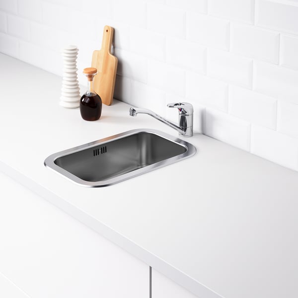 Stainless steel kitchen sink with tap and accessories, against white tiled wall.