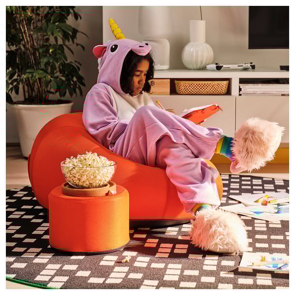 Person in unicorn onesie sits in orange inflatable chair, reading a book while eating popcorn. Colourful rug and toys surround.