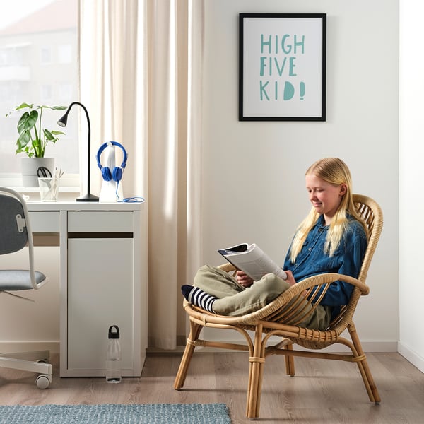 Child reading in wicker armchair by desk with laptop, lamp, and headphones.