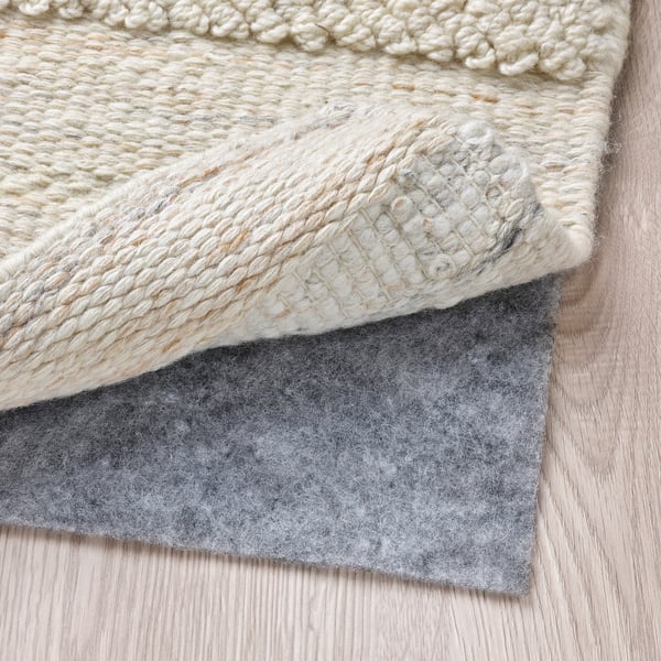 A close-up of a beige woven rug laid on a light wooden floor, with grey felt underlay visible, showcasing its texture and functional placement.