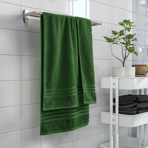 Green BROKGLIM towel hangs large on wall rack, near plant & folded dark towels on white stand.