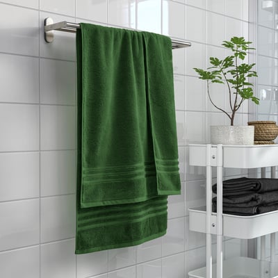 Green BROKGLIM towel hangs large on wall rack, near plant & folded dark towels on white stand.