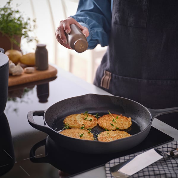 Person seasoning fried food in cast iron skillet with pepper grinder.