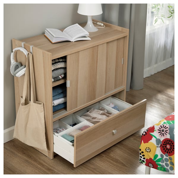 Light wood storage cabinet with open drawer & door, headphones, books, lamp.