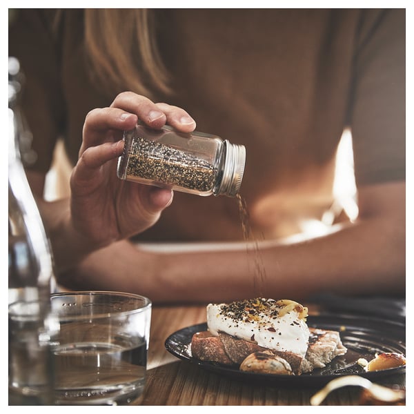 A person sprinkles pepper from a CITRONHAJ glass shaker onto food on a black plate, with a clear glass cup beside it.