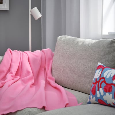 Cosy grey sofa with bright pink DAGGKÅPOR fleece blanket and floral pillow. Soft, recycled polyester, curtains casually over armrest.