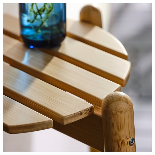 A close-up of a wooden bench with smooth, polished slats. A blurred vase is seen in the background.
