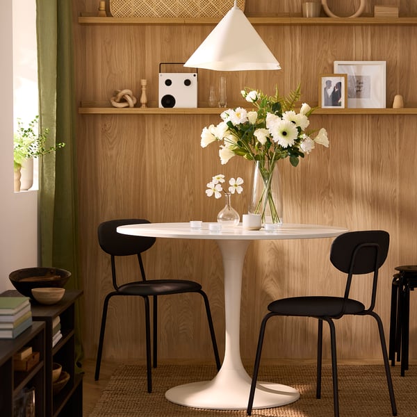 Modern, white DOCKSTA round dining table with black chairs, marble-like top, surrounded by a wooden wall with shelves and a hanging lamp.