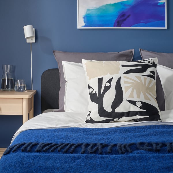 Blue bedroom with floral bed, grey pillows, blue throw, wooden bedside table, glass carafe, art.