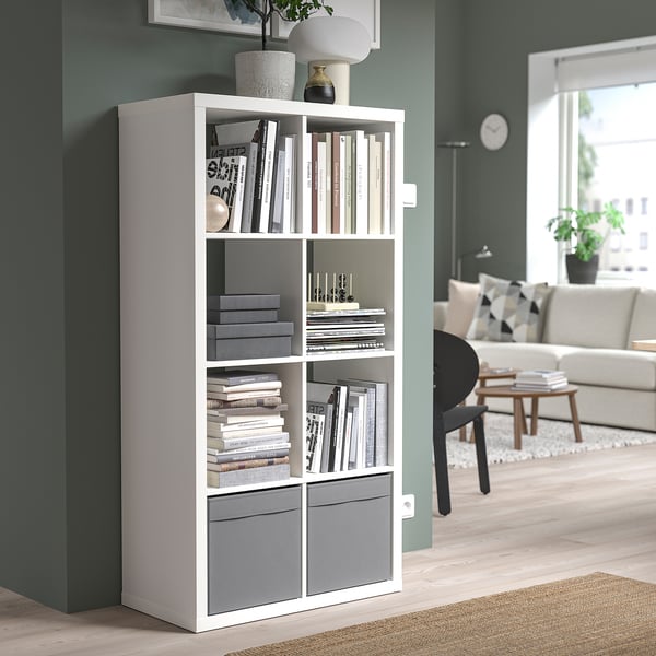 White DRÖNA storage box on tall shelf, containing books, next to living room with sofa and table.