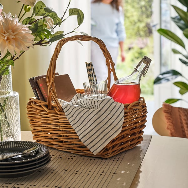 Wicker basket with handle containing lingonberry syrup, cups, napkins, and plates on wooden table.