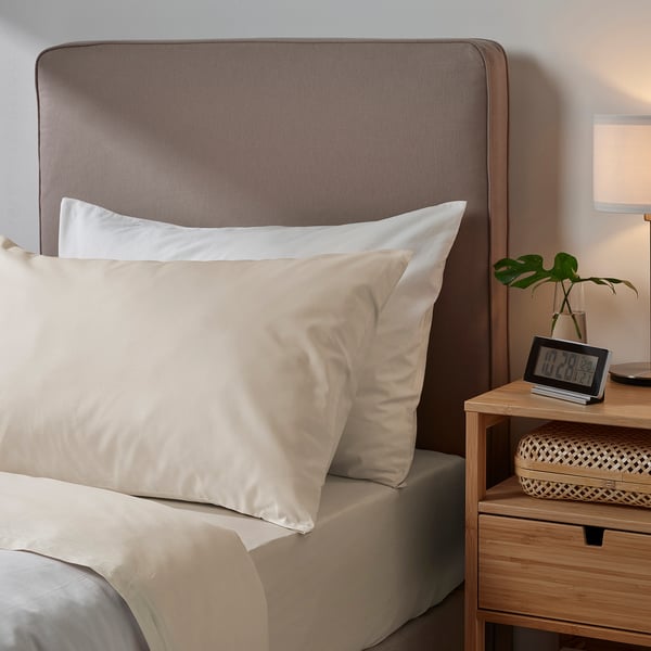 Bedroom scene with light-colored DVALA bedding. Pillowcase and sheets are 100% cotton. Wooden headboard and nightstand, clock and plant nearby.
